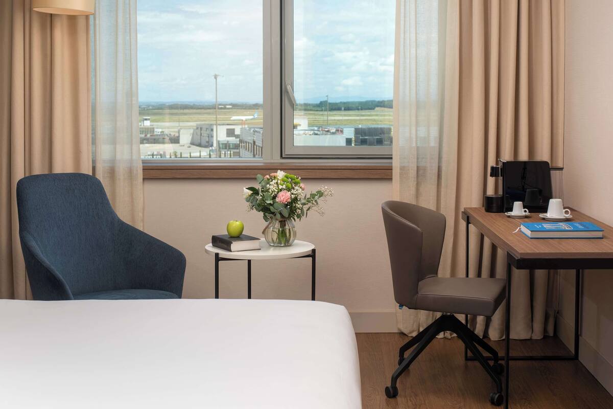 Ein Hotelzimmer mit einem Bett, zwei Stühlen, einem kleinen Tisch mit Blumen, einem Buch, einer Kaffeemaschine und Blick auf einen Flughafen.