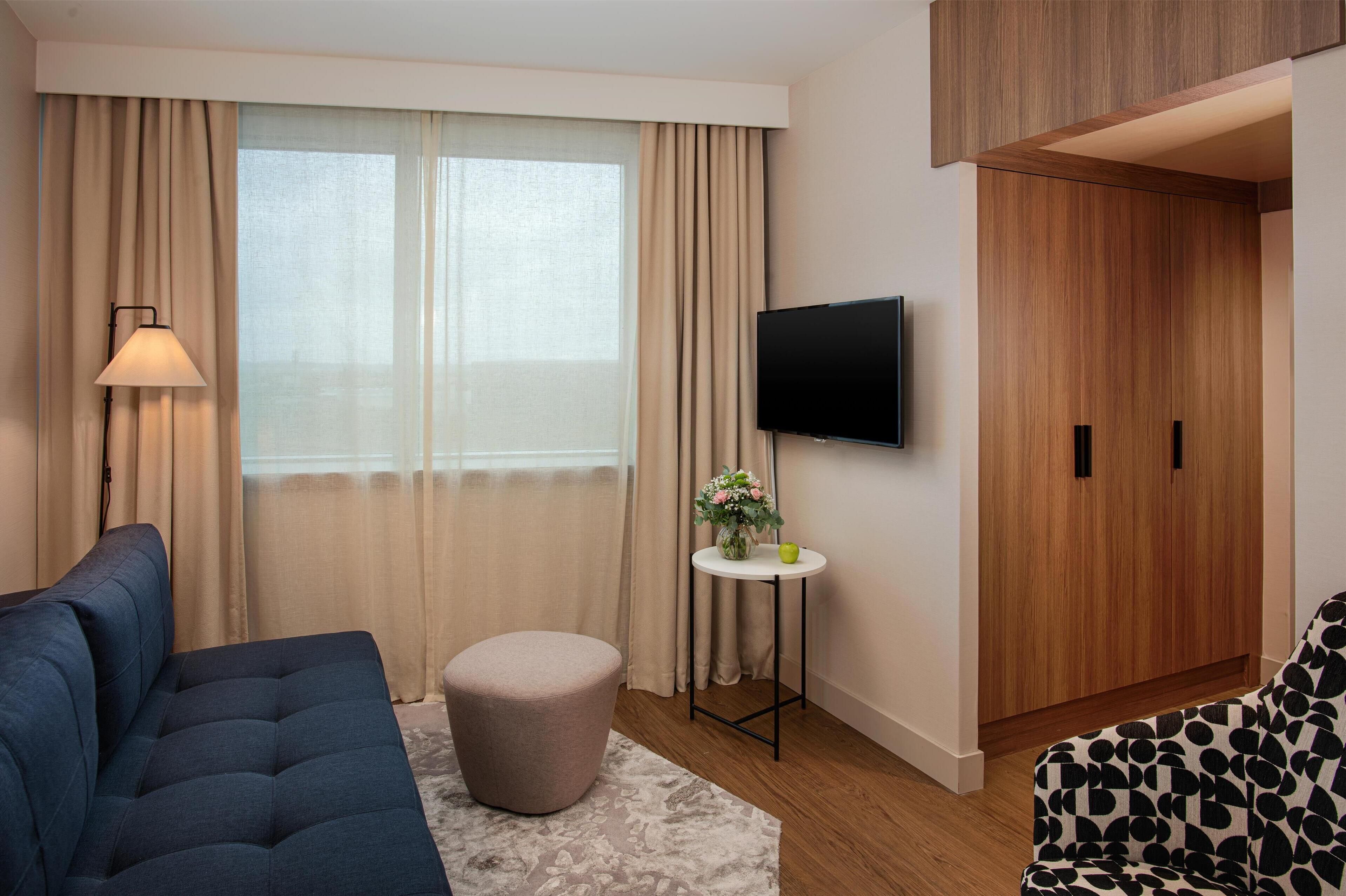 Ein modernes Hotelzimmer mit einer Couch, Hocker, Fernseher und einem Holzschrank.