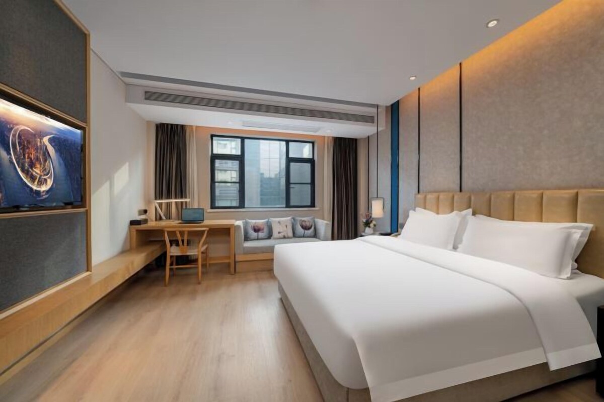 A modern hotel room with a large bed, a desk with a chair, a TV mounted on the wall, and a window providing a city view.