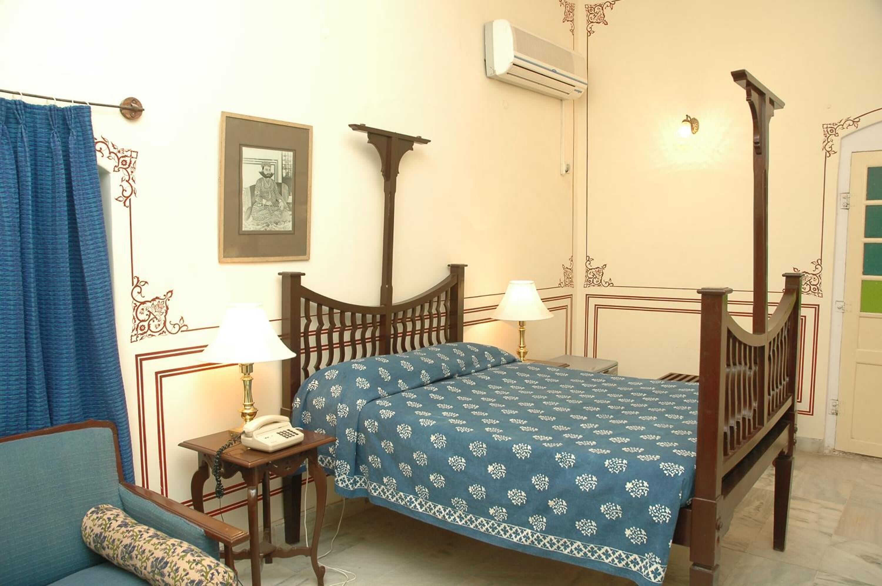 A traditional four-poster bed with a blue patterned bedspread, a bedside table with a lamp, a framed picture on the wall, and a blue curtain.