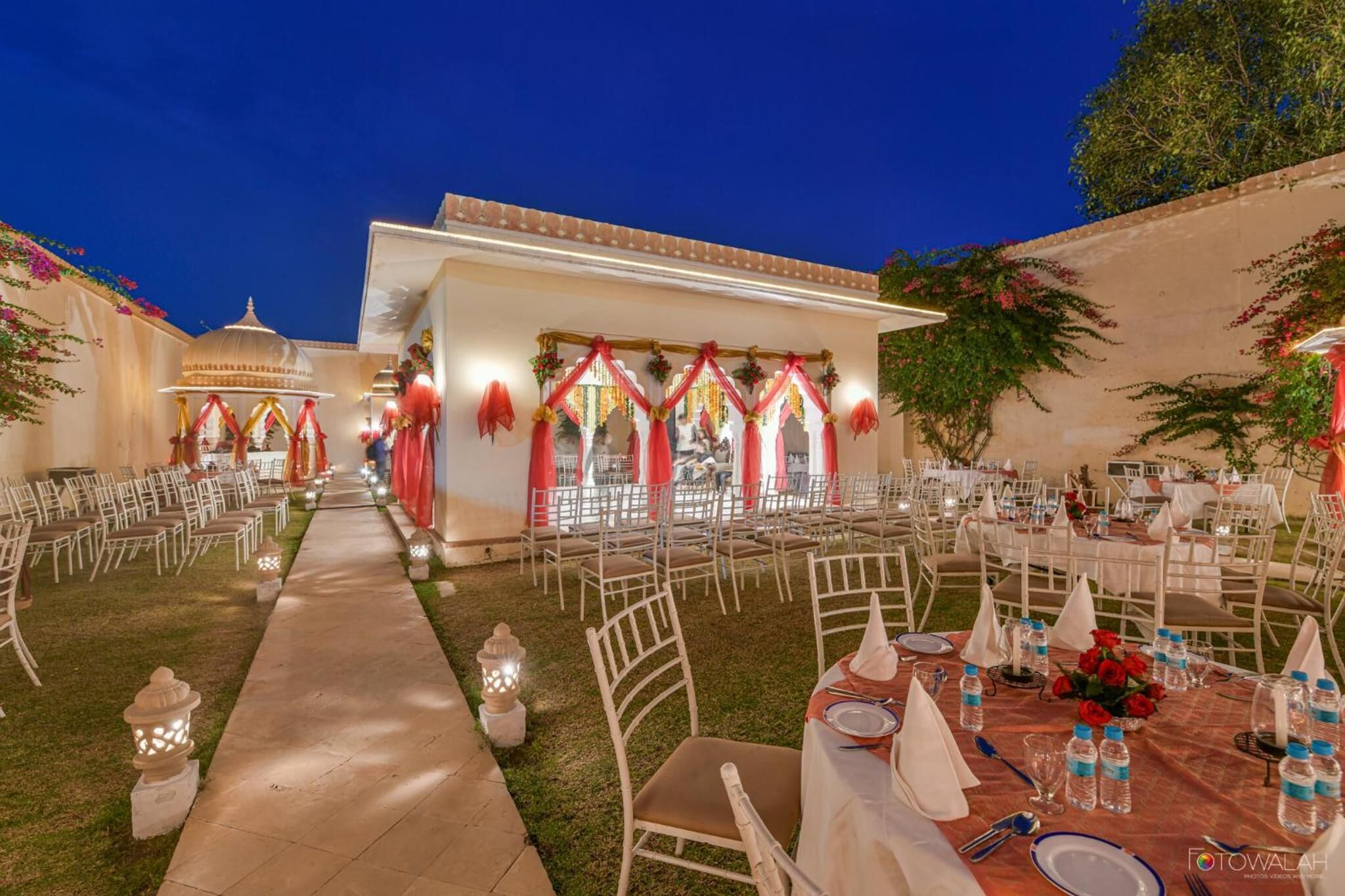 Un espace événementiel en plein air, avec des rangées de chaises et de tables blanches disposées pour les repas, décoré de draperies rouges et d’arrangements floraux.