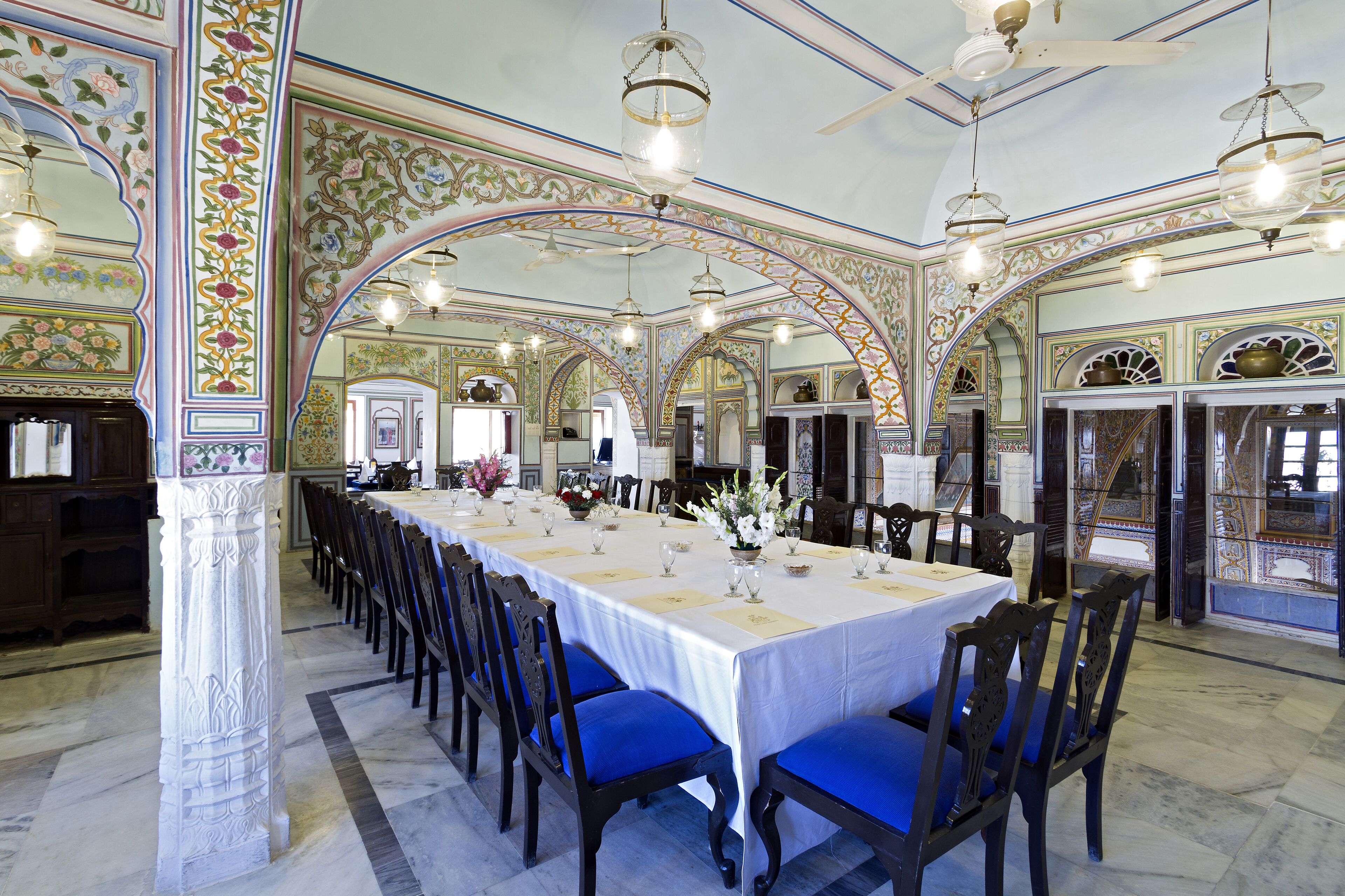 Une longue table à manger, des chaises bleues, un plafond décoré et des lustres.