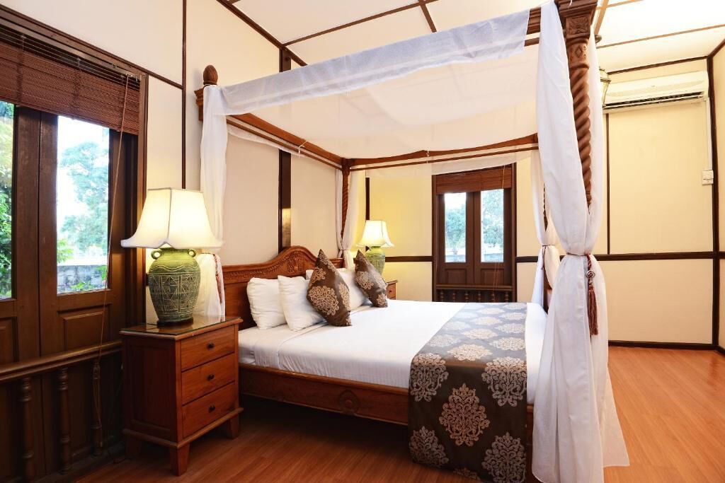 A four-poster canopy bed with a white bedspread and pillows, a nightstand with a lamp, and a window with a view of greenery.