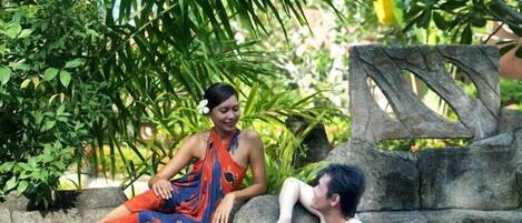 A person in a red and blue outfit sitting on a stone ledge by a pool with water lilies, surrounded by lush greenery.