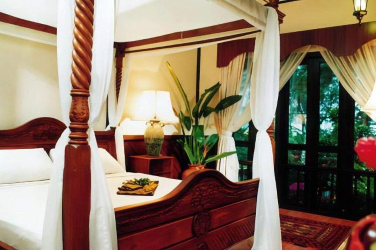 A four-poster bed with a canopy, a bedside table with a lamp, a potted plant, and a view of the outdoors through a window with curtains.