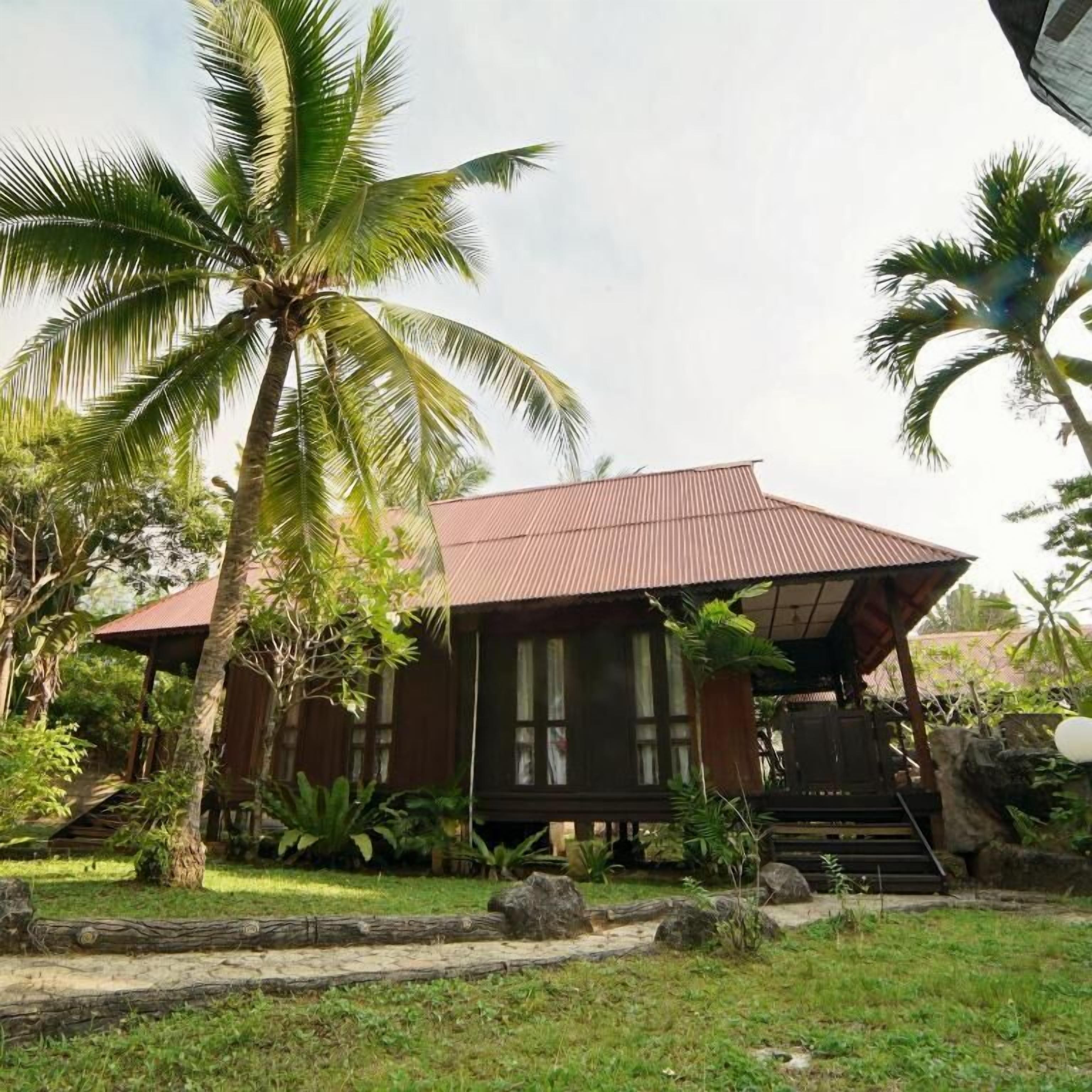 A wooden house with a sloped roof, surrounded by palm trees and greenery.