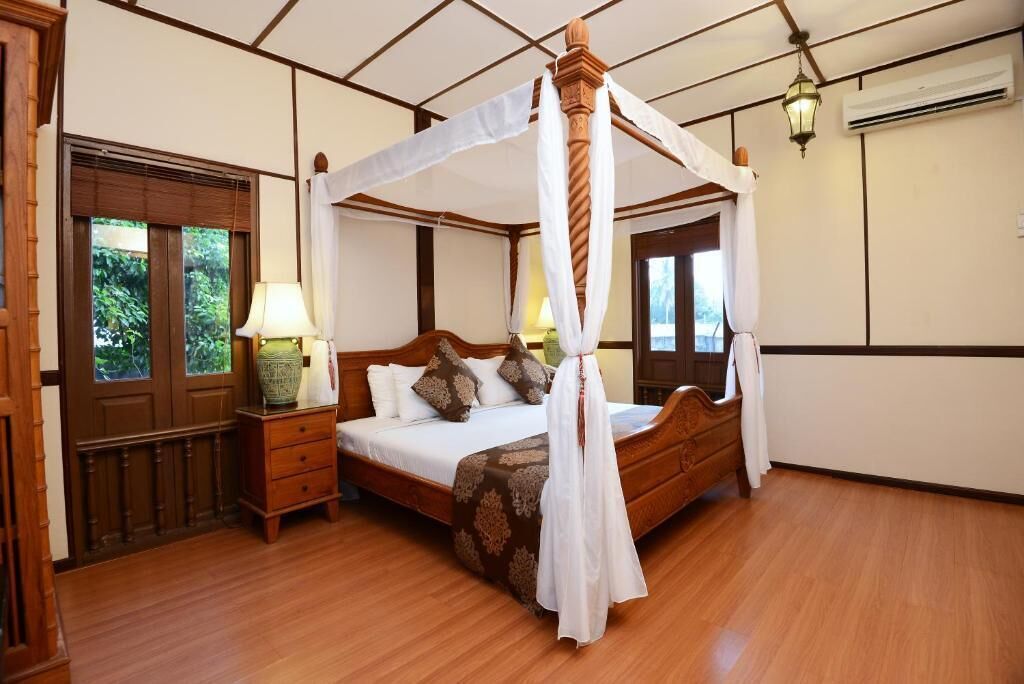 A four-poster canopy bed with a wooden frame and white drapes, a bedside table with a lamp, and a view of greenery through the window.