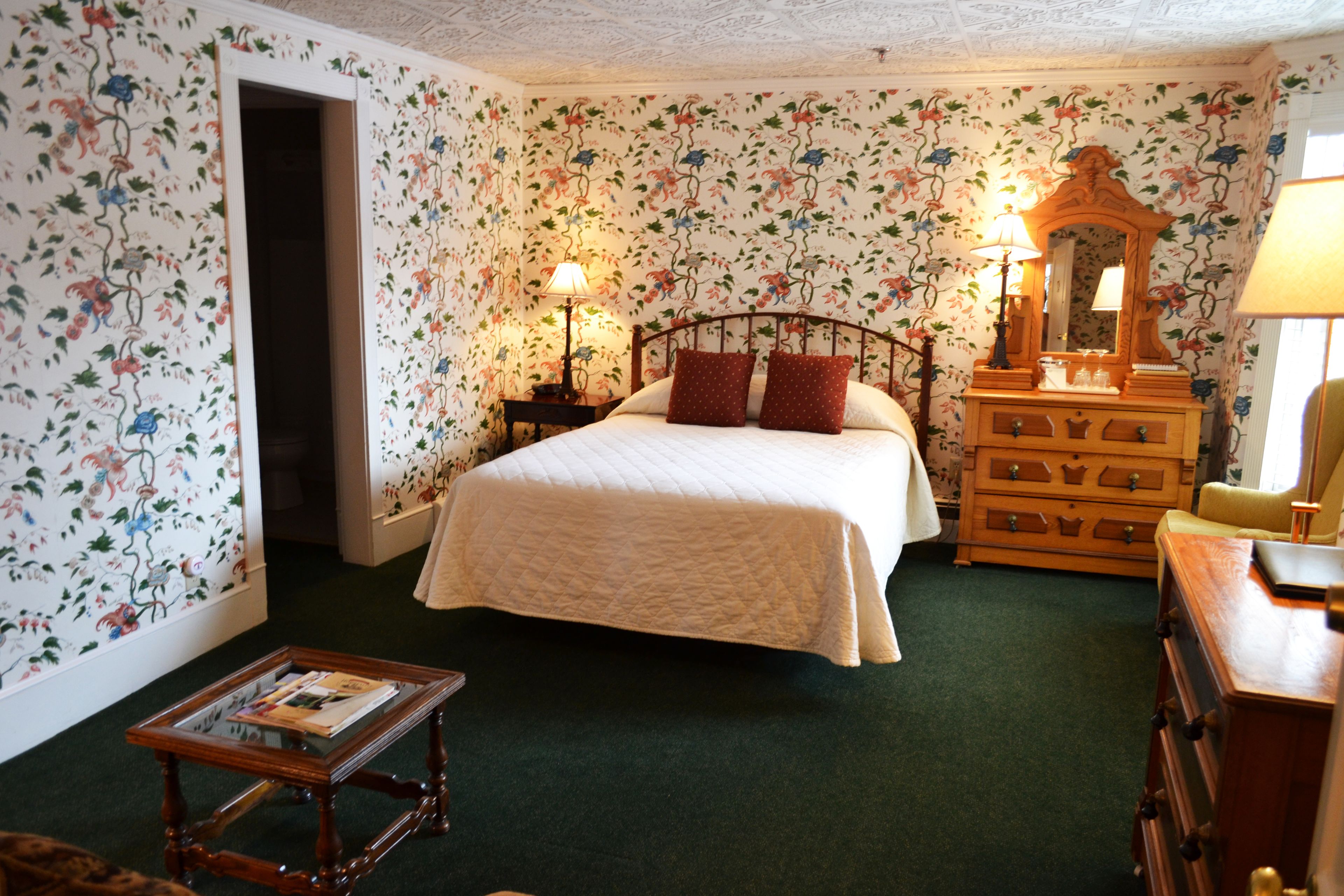 A bedroom with floral wallpaper, a bed with red pillows, a dresser, a mirror, and a nightstand with a lamp.
