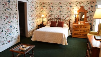 A bedroom with floral wallpaper, a bed with red pillows, a dresser, a mirror, and a nightstand with a lamp.