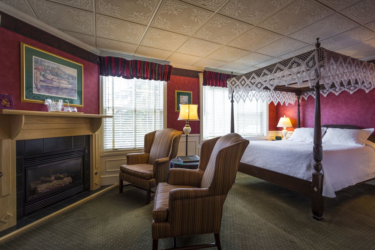 A bedroom with a four-poster bed, a fireplace, two armchairs, and a nightstand with a lamp.