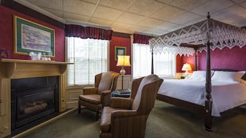 A bedroom with a four-poster bed, a fireplace, two armchairs, and a nightstand with a lamp.