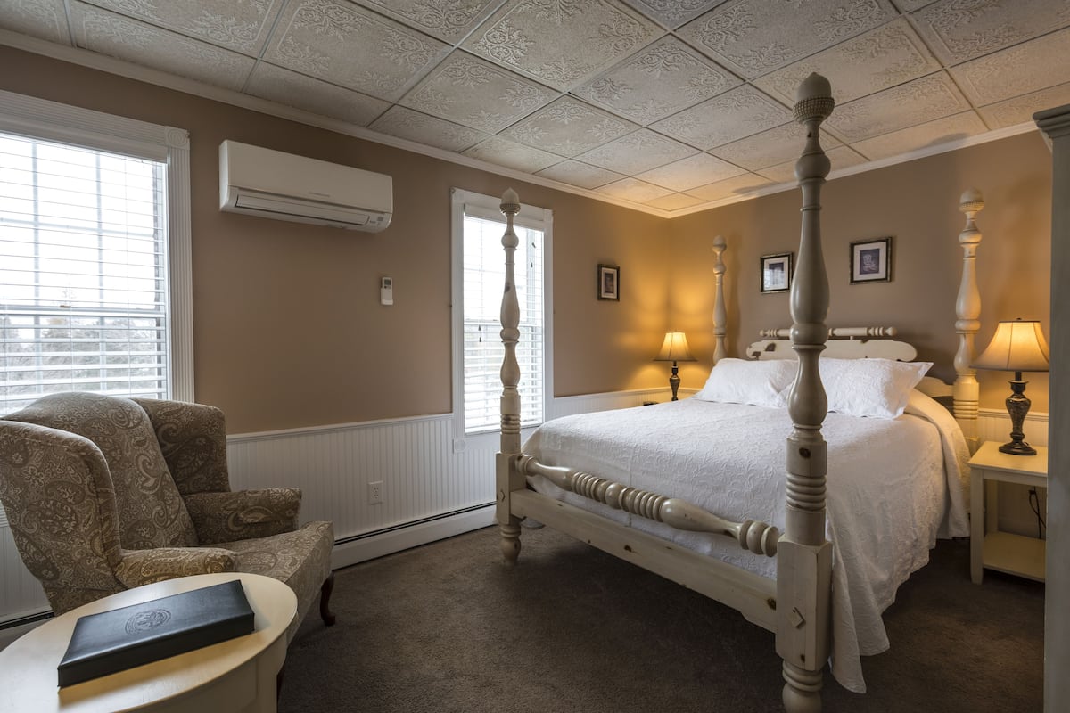 A four-poster bed with a white bedspread, a chair with a patterned upholstery, and a bedside table with a lamp.