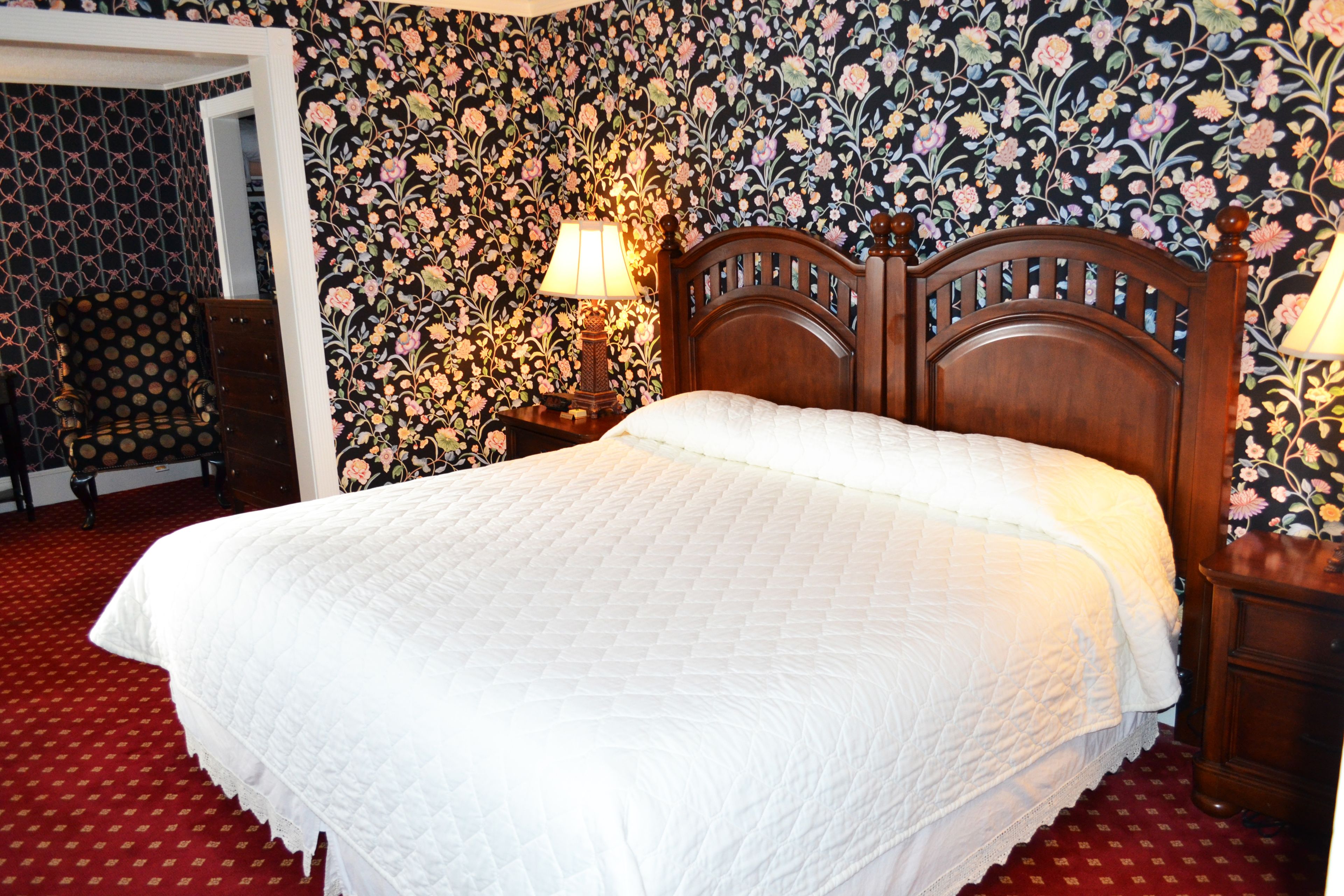 A neatly made bed with a white quilt in a room with floral wallpaper and a red carpet.