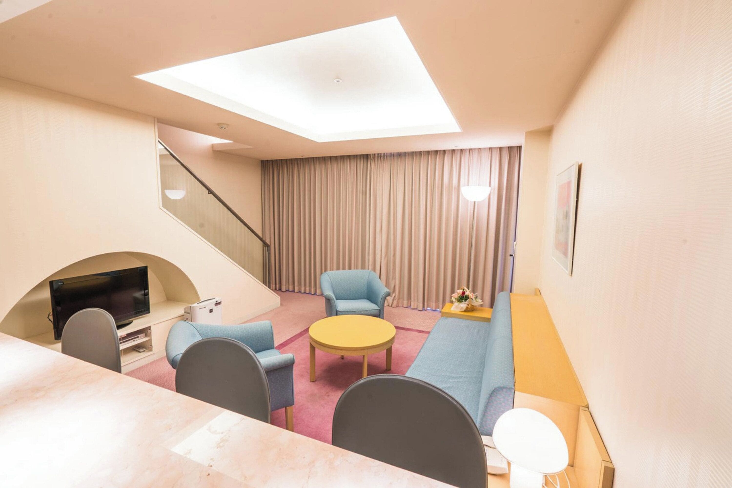 A modern hotel room with a flat-screen TV, a sofa, a round coffee table, and a staircase.