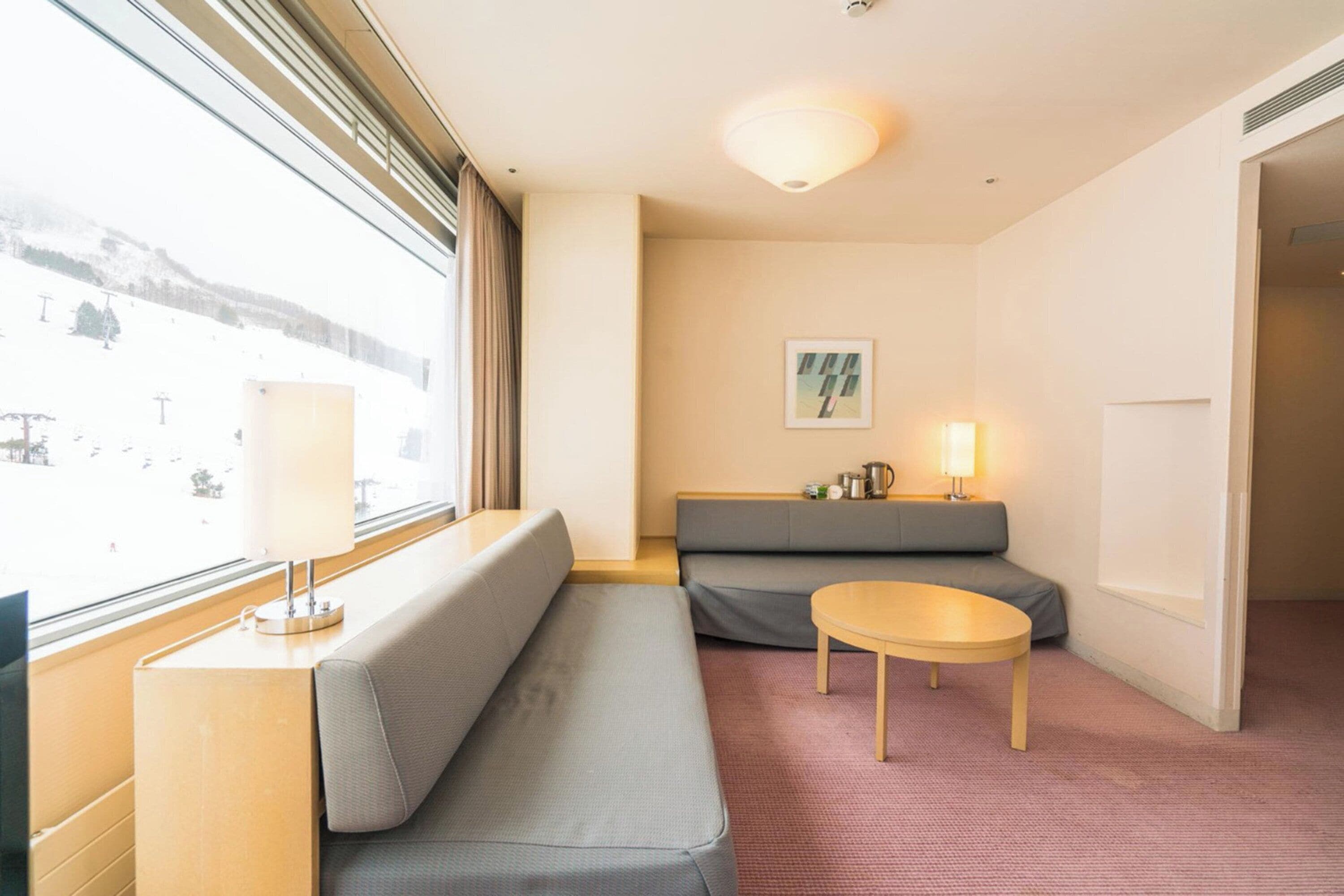 A hotel room with a sofa, a small table, a lamp, and a view of a snowy landscape through the window.