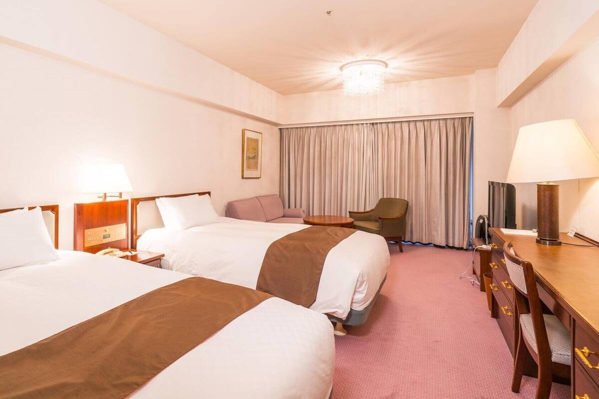 A hotel room with two beds, a desk, a chair, a television, and a large window with curtains.