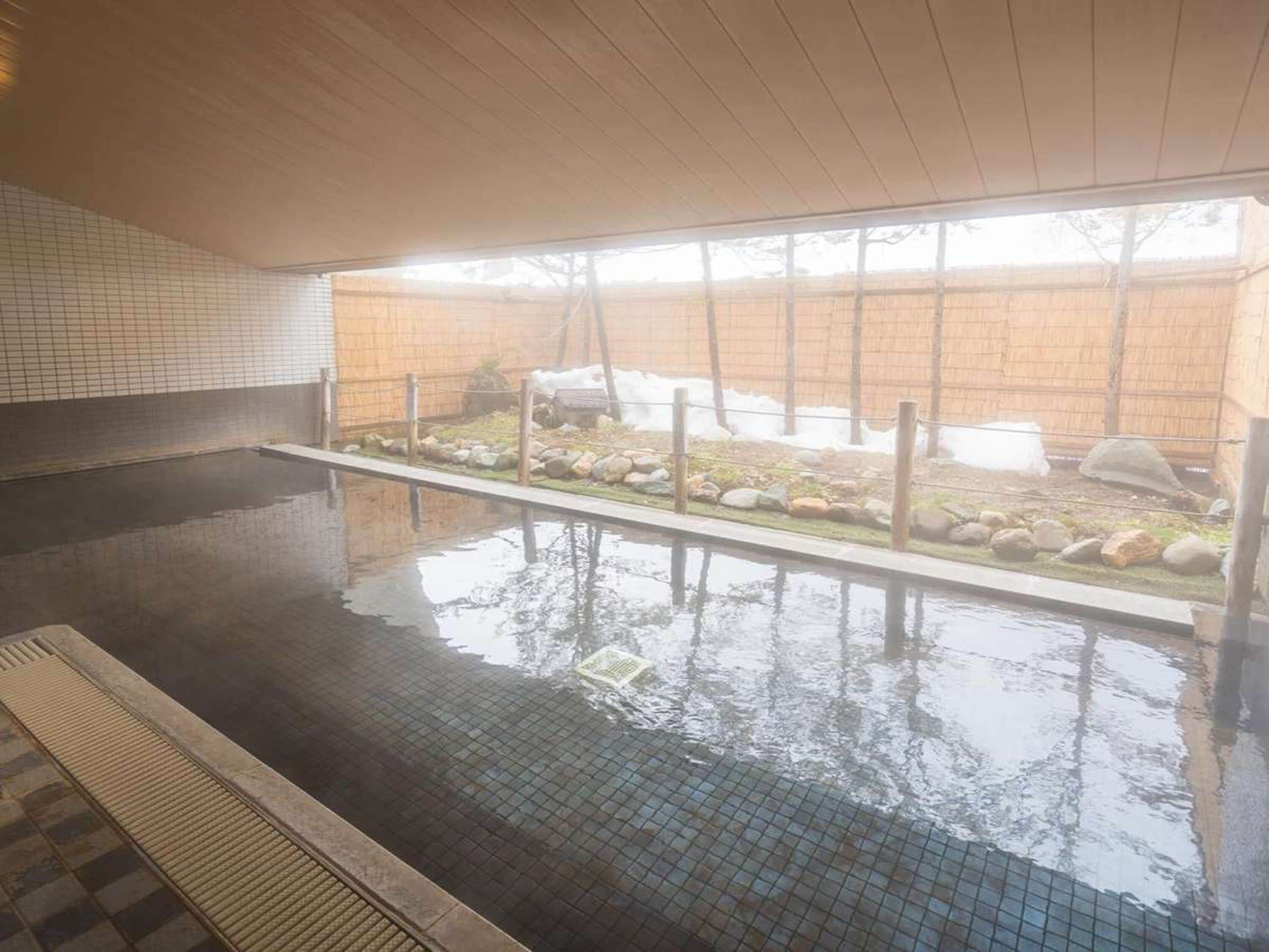 Una piscina interior con agua cristalina, suelo de baldosas y vistas a un área exterior vallada con rocas y una valla de madera.