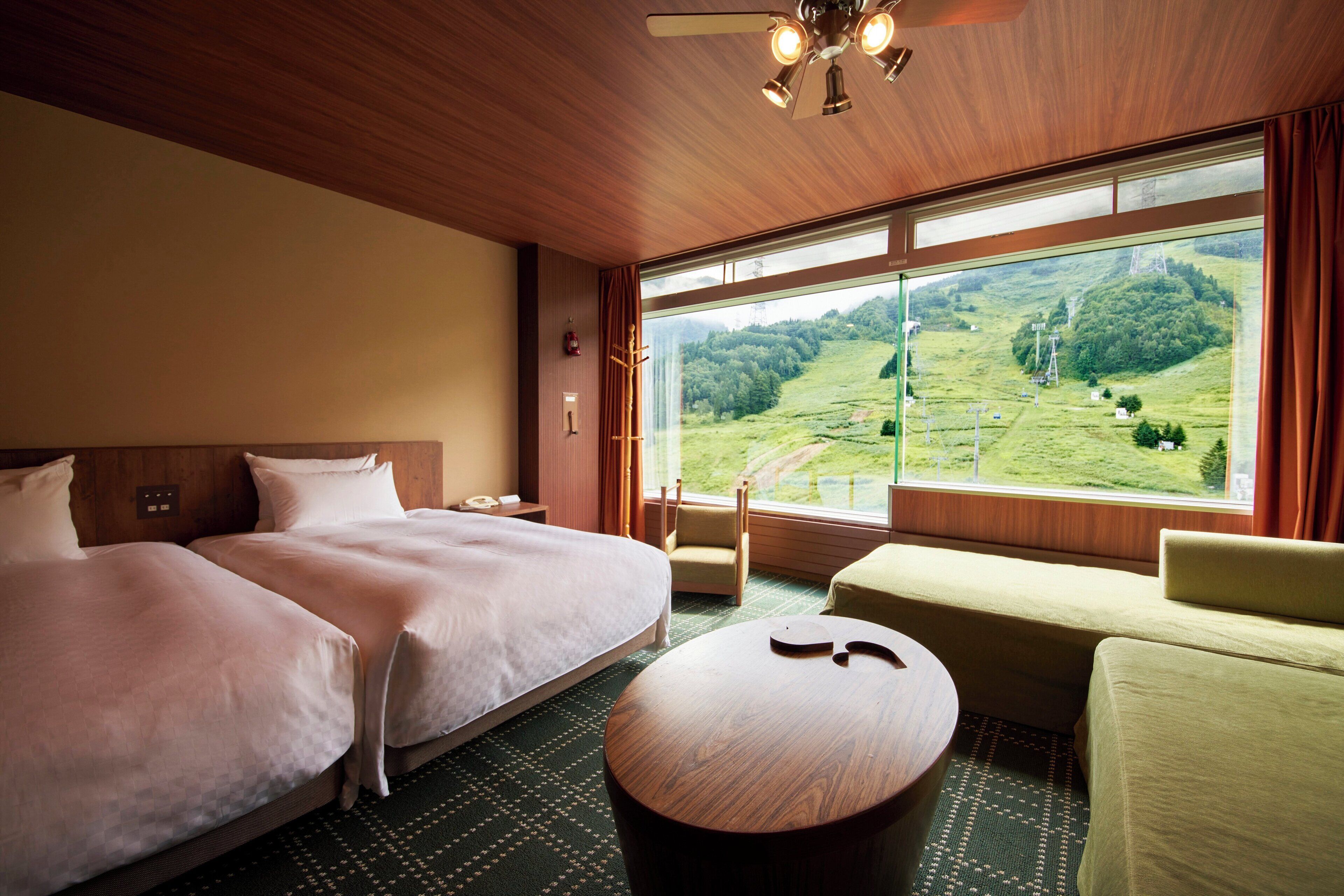 A hotel room with two beds, a sofa, a round table, and a large window offering a view of a green landscape.
