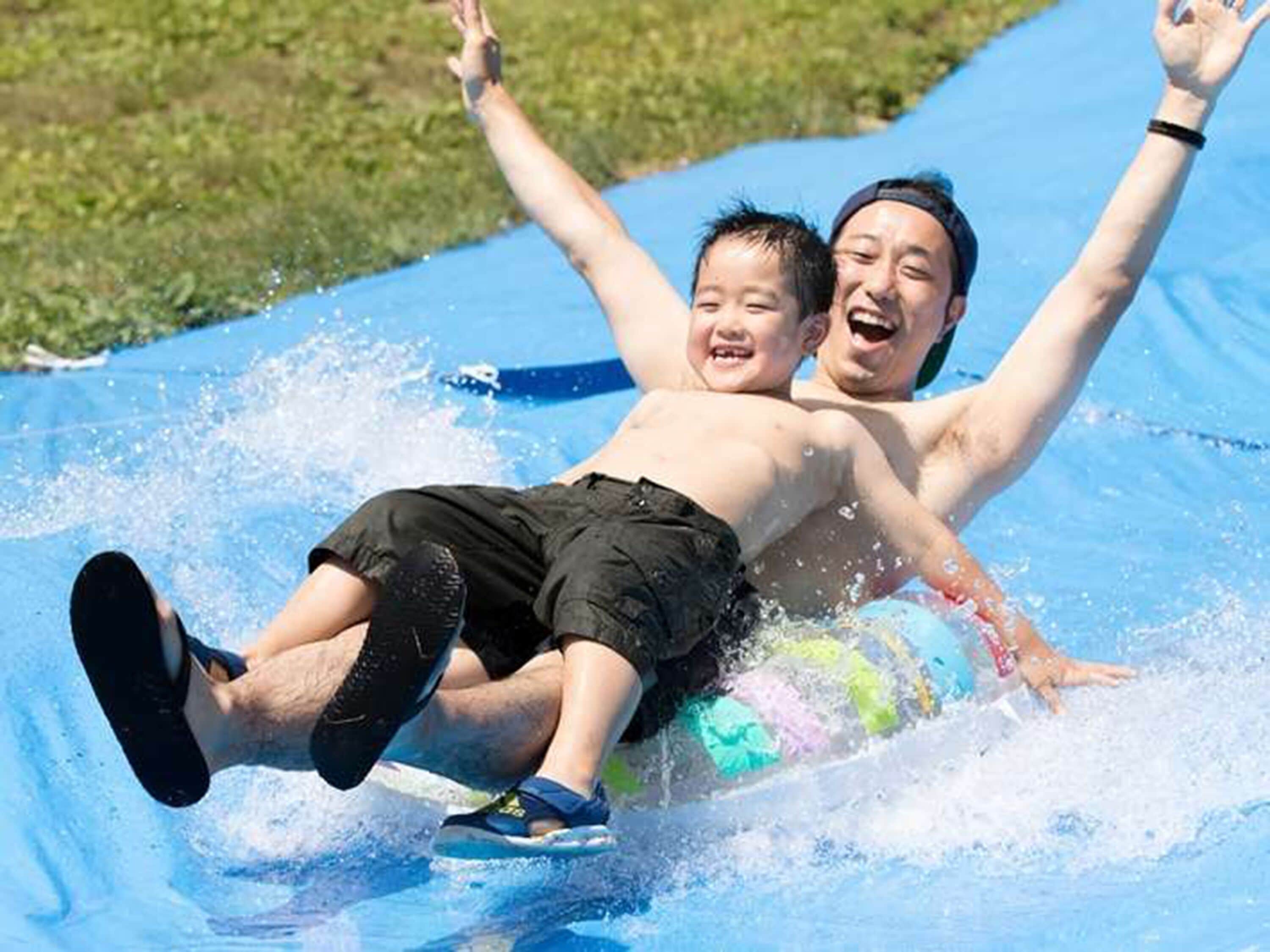 Une personne allongée sur un toboggan gonflable bleu, les bras levés, vêtue d’un short noir et de sandales.