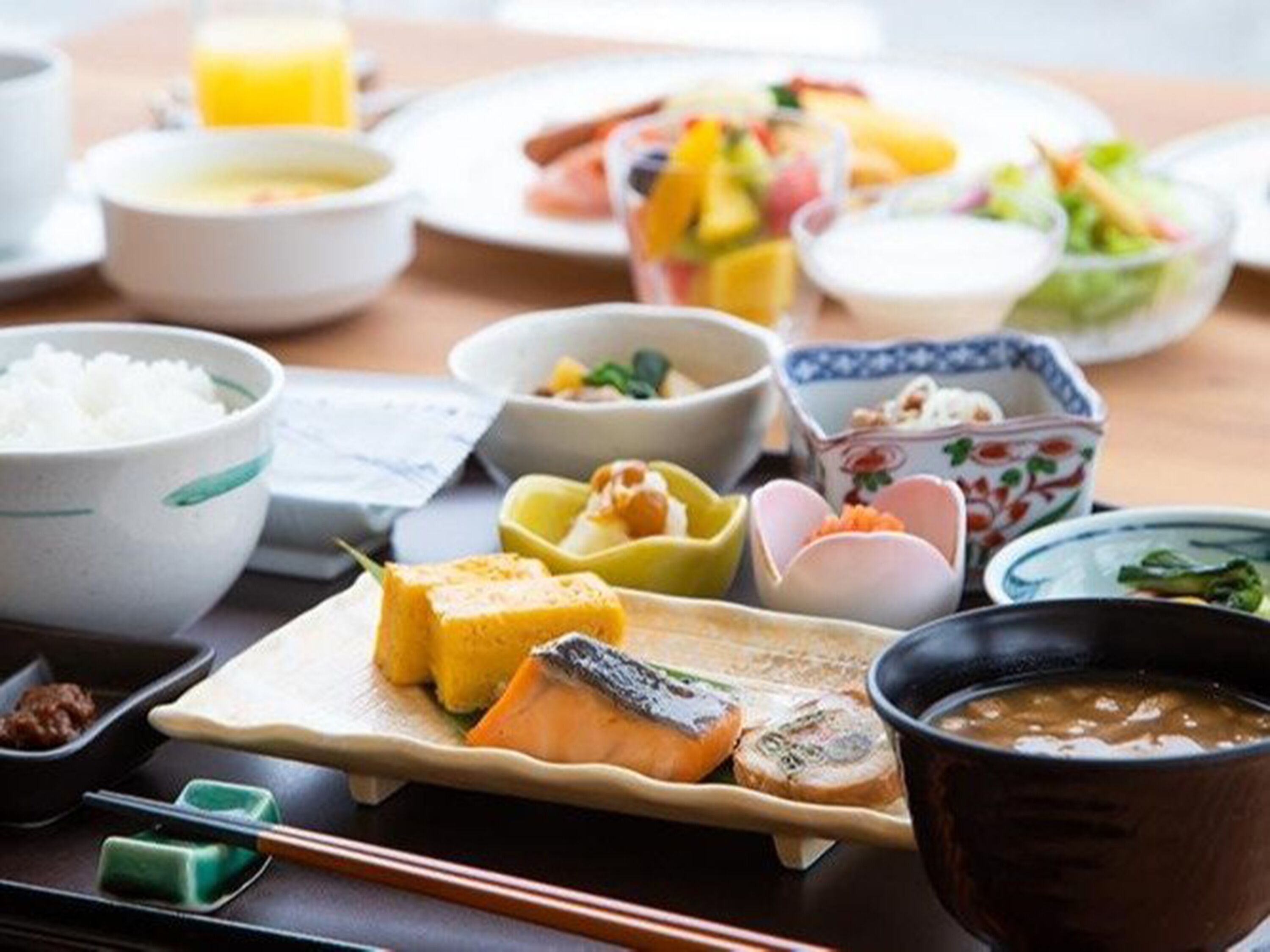 Különféle japán ételeket kínálunk, köztük rizst, halat és zöldségeket, melyeket egy fa asztalon szolgálunk fel.