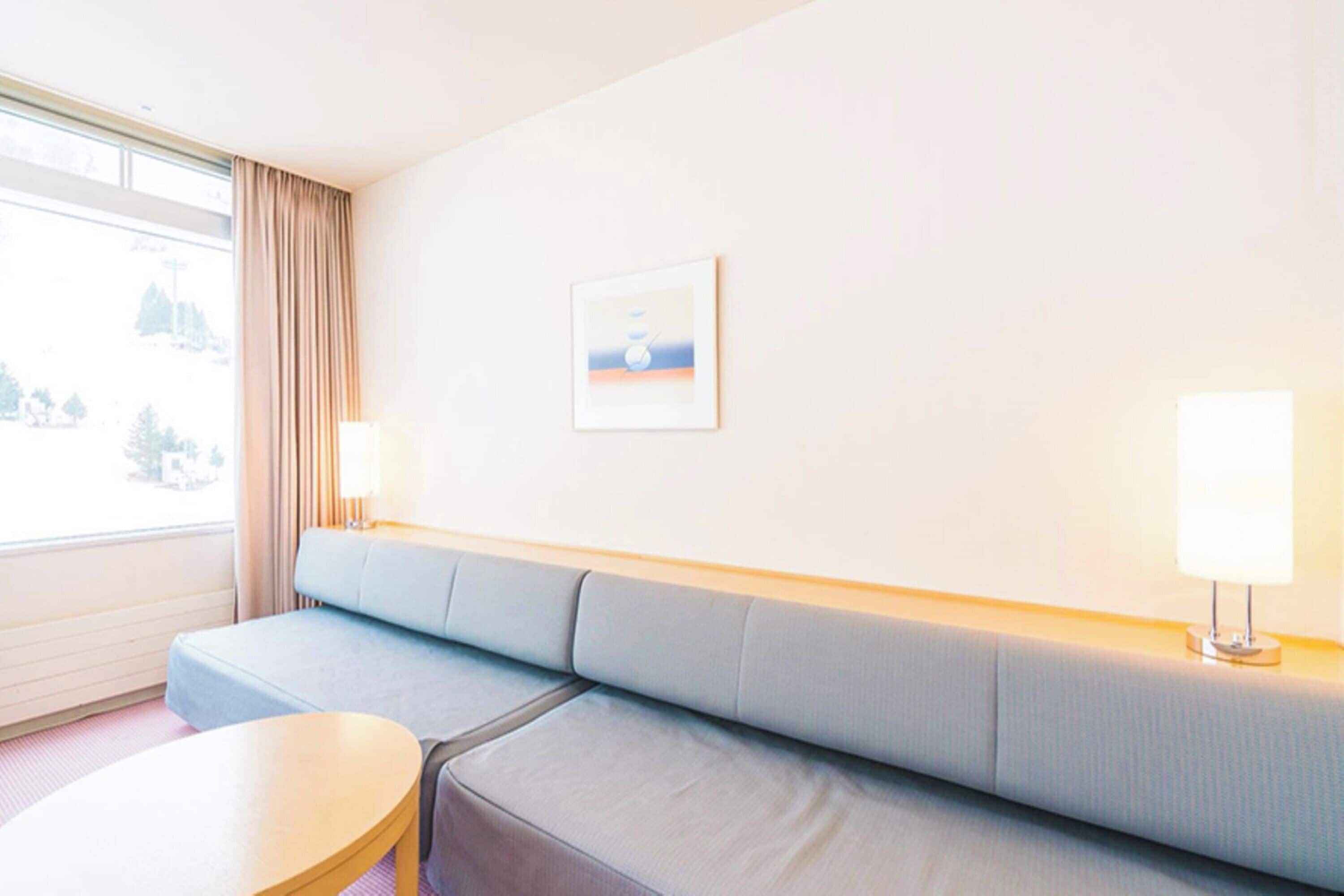 A hotel room with a light grey sofa, a round wooden table, and a window with curtains.