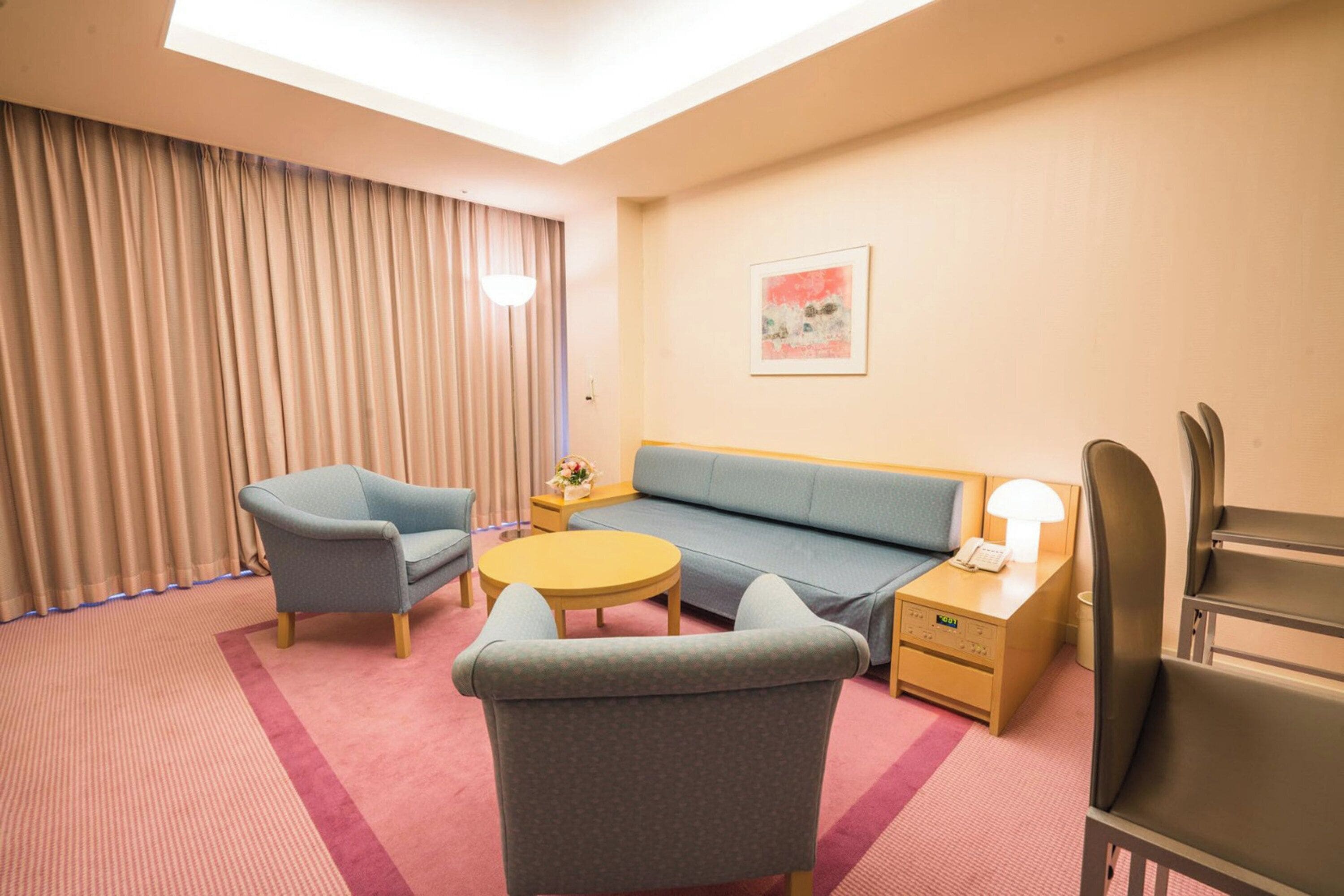 A hotel room with a sofa, two armchairs, a small round table, a bedside table with a lamp, and a painting on the wall.