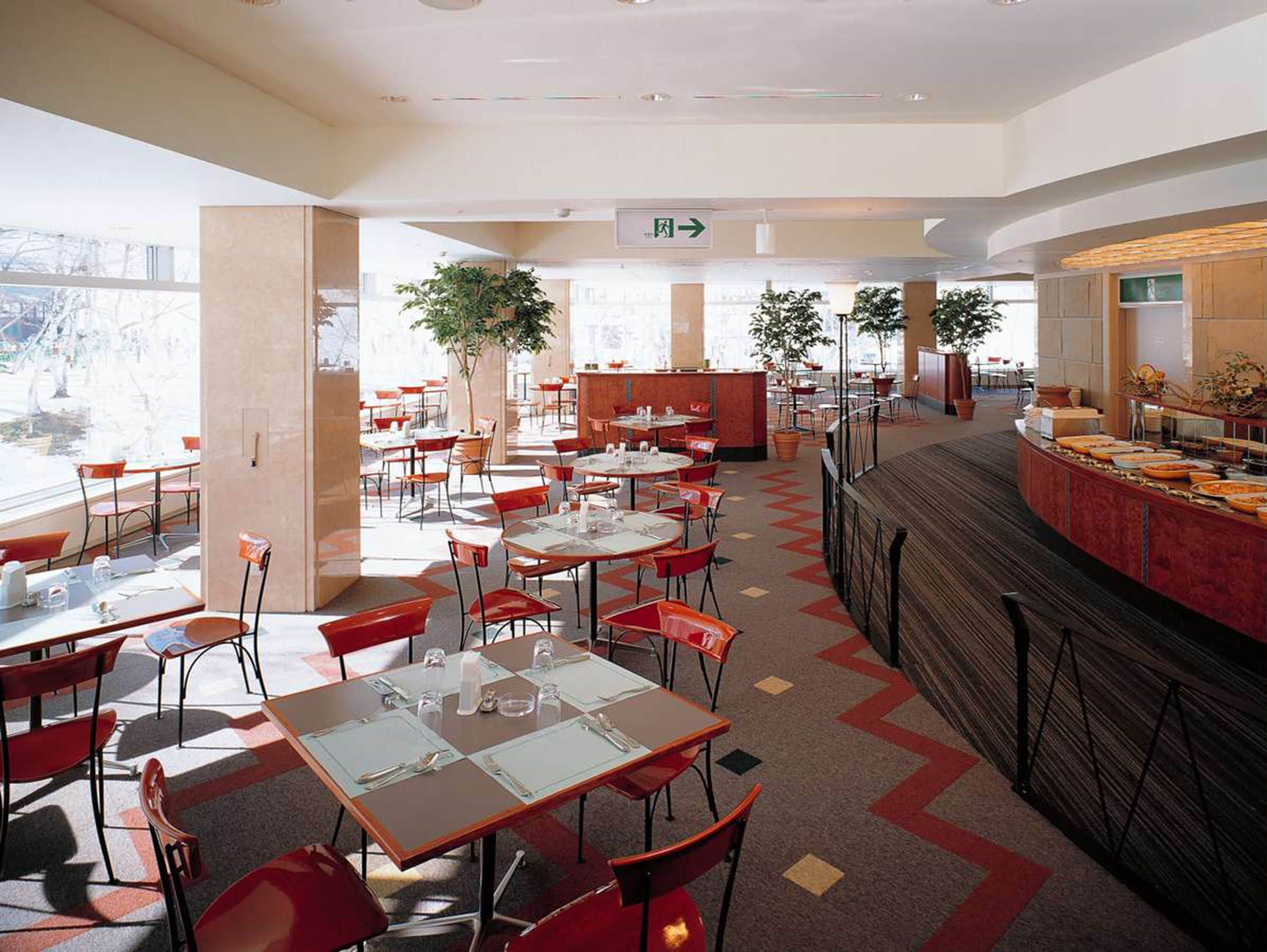 A hotel dining area with tables and chairs, a buffet counter, and potted plants.
