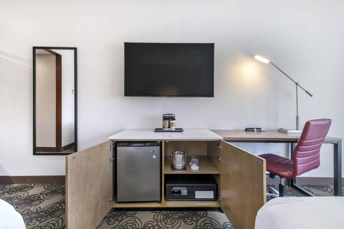 Habitación de hotel con cama, escritorio con silla, refrigerador, cafetera, espejo y televisor en la pared.