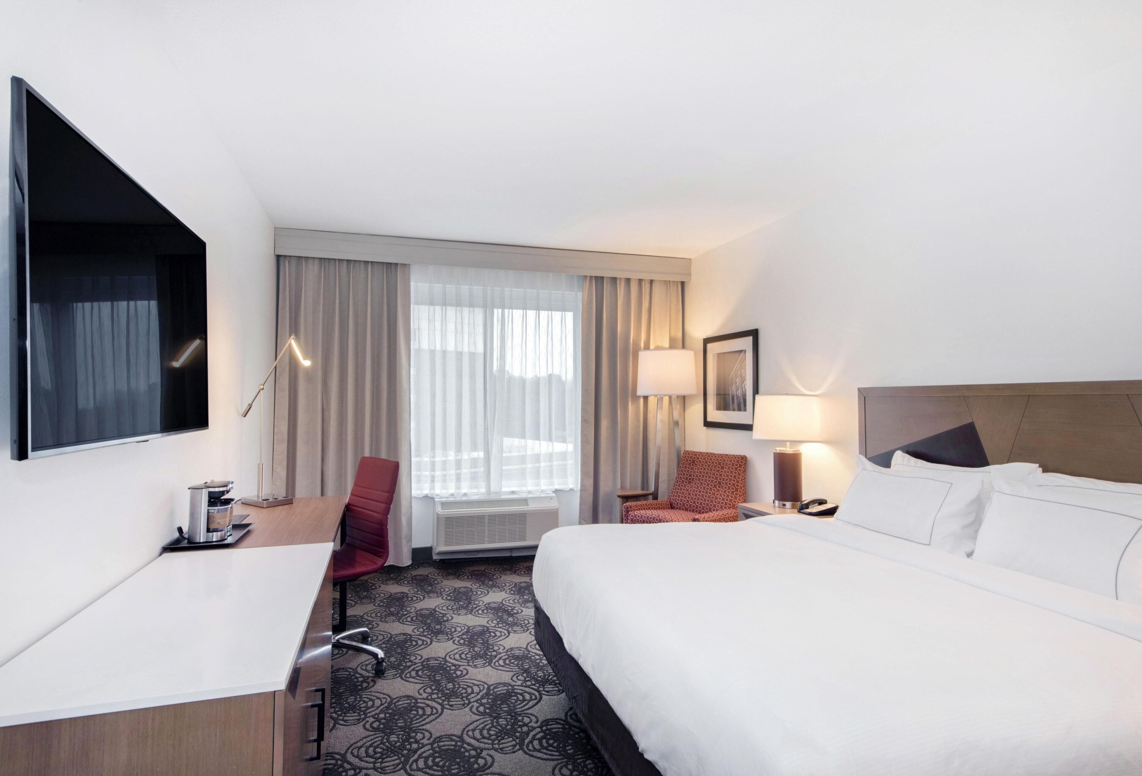 A hotel room with a bed, desk, chair, and a large window with curtains.