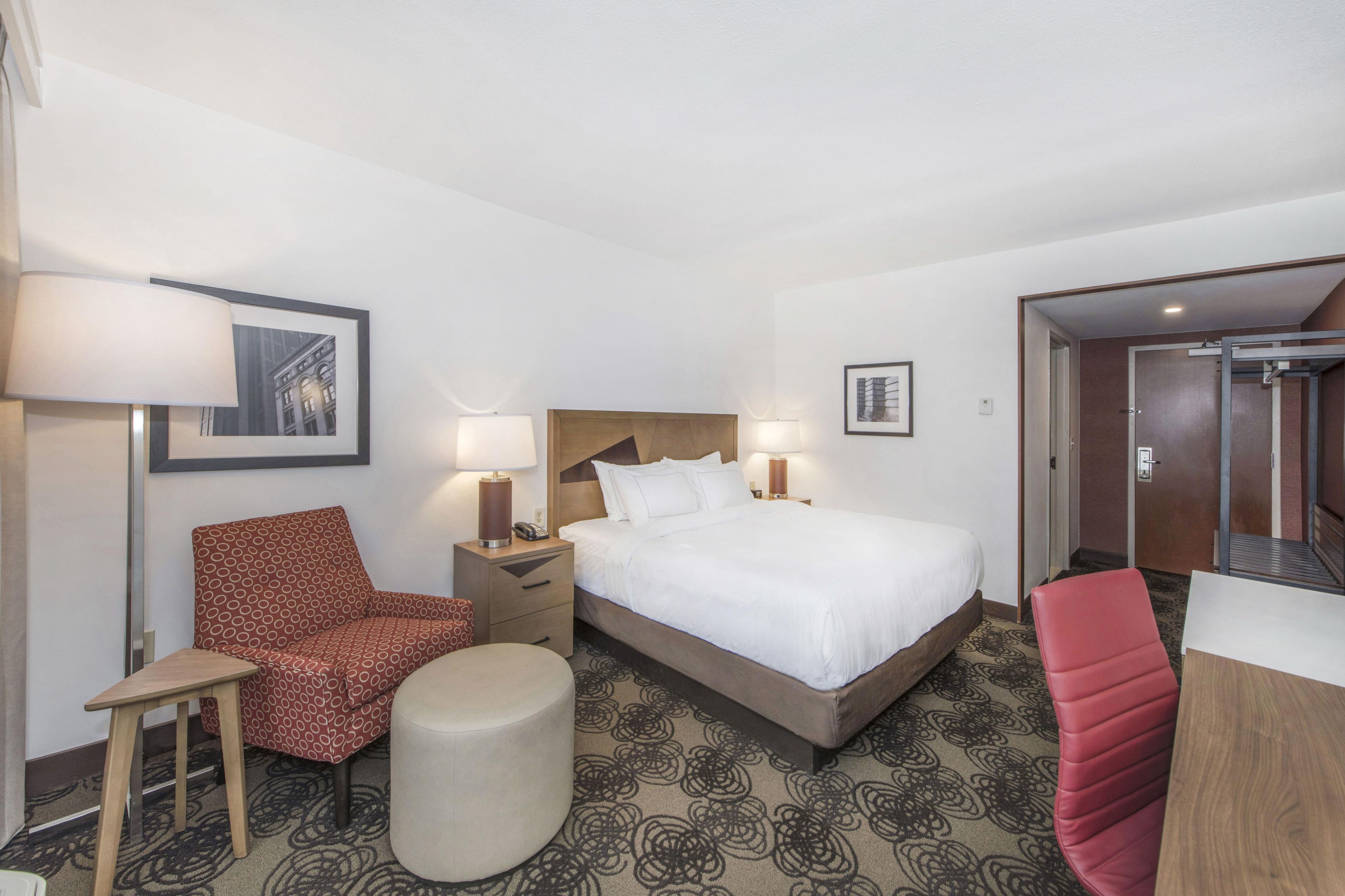 Een hotelkamer met een bed, een stoel, een tafeltje, een lamp en een foto aan de muur.