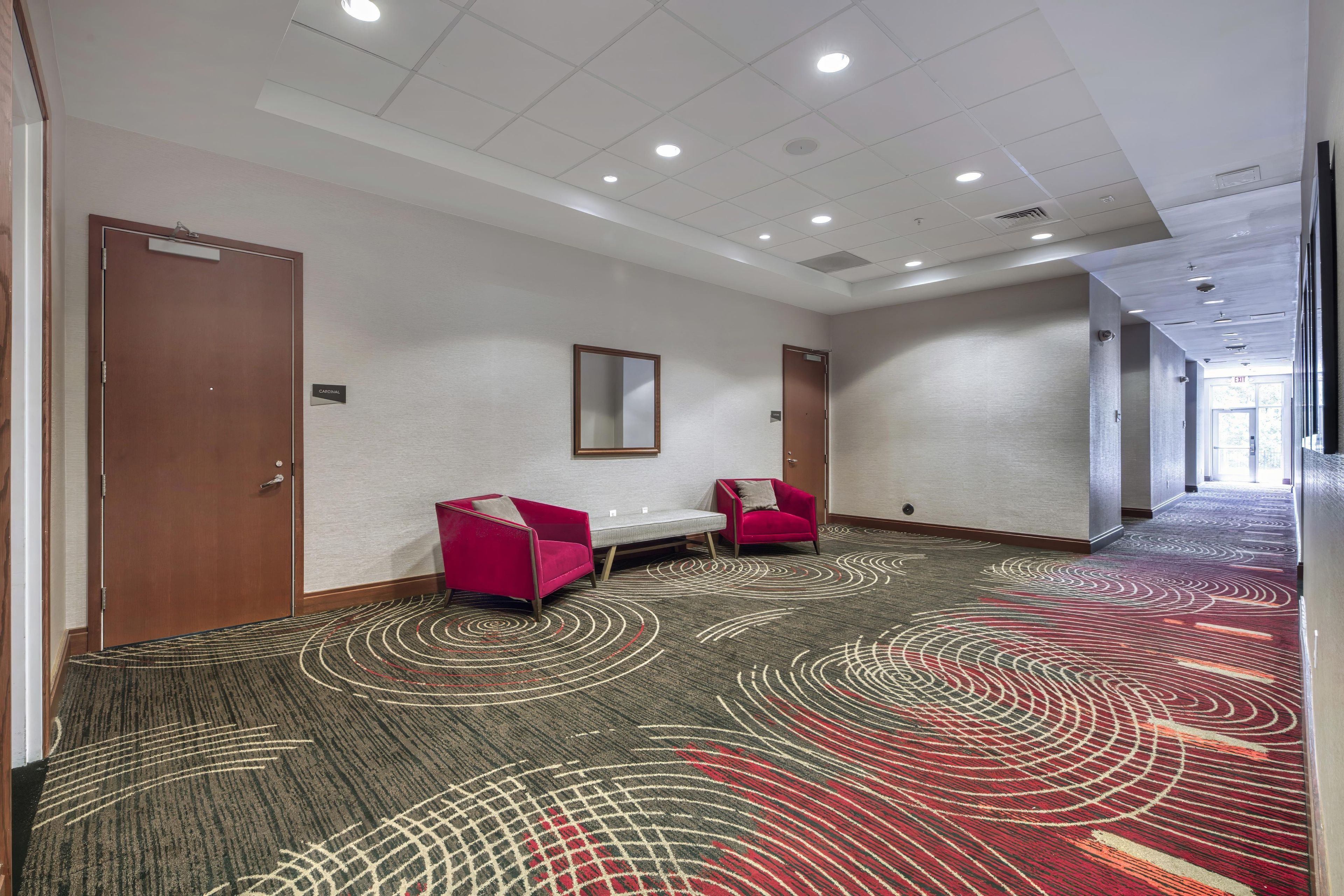A hotel corridor with a patterned carpet, two red armchairs, a white table, and a mirror on the wall.
