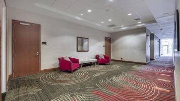 A hotel corridor with a patterned carpet, two red armchairs, a white table, and a mirror on the wall.