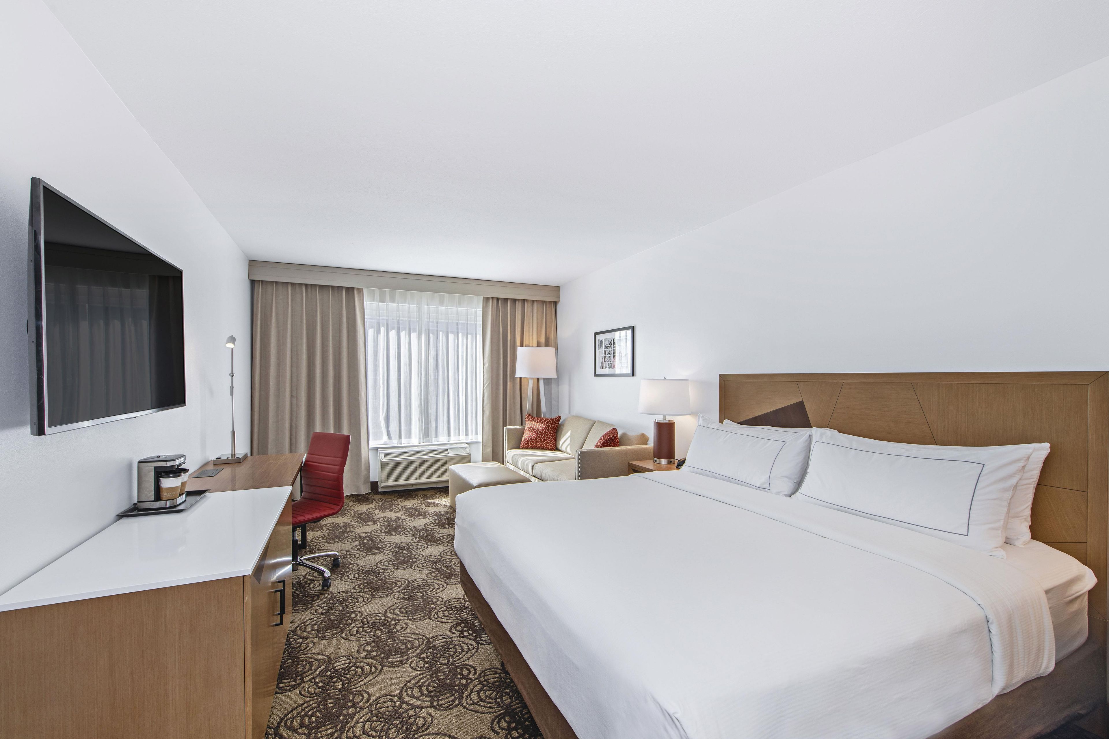 A hotel room with a bed, a desk, a chair, a TV, a window with curtains, and a carpet.