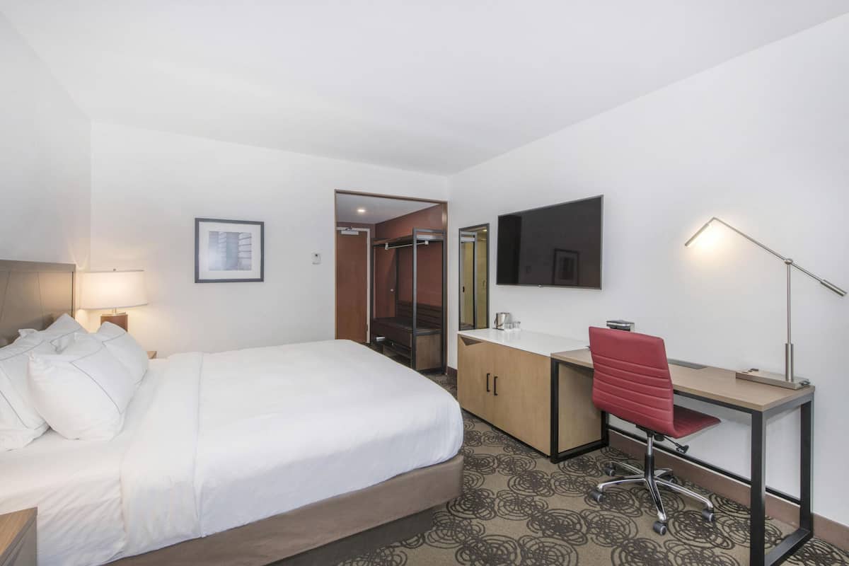 Habitación de hotel con cama, escritorio, silla, espejo, lámpara y puerta de baño.