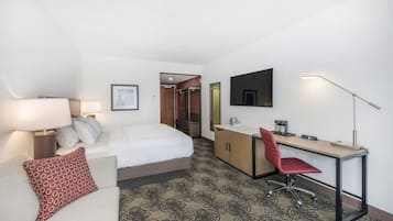 A hotel room with a bed, a desk, a chair, a desk lamp, a TV, and a patterned carpet.