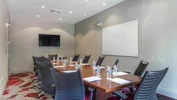 Un salón de conferencias con una mesa larga, sillas, botellas de agua, una pizarra blanca y un televisor en la pared.