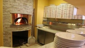 A stone fireplace with a wood-burning stove, a metal rack with pizza boxes, and a stack of plates. - Hotel Bernina (Tirano)