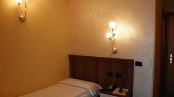 Une petite chambre avec un lit simple, une tête de lit en bois, une table de chevet avec un téléphone et une télécommande, et des luminaires fixés au mur.