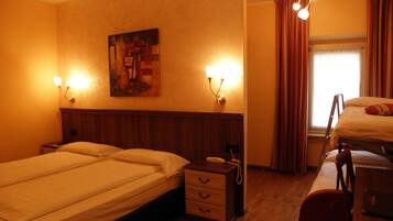 A hotel room with two beds, a wooden headboard, a bedside table with a lamp, a painting on the wall, and a window with curtains.
