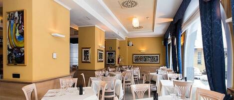 Un restaurante con varias mesas y sillas, una pared amarilla con obras de arte enmarcadas y un amplio ventanal con cortinas.