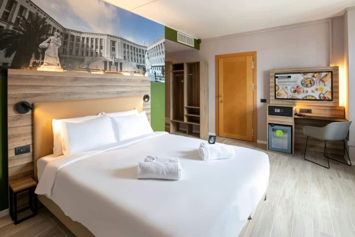 A hotel room with a large bed, a desk, a chair, a television, and a mural on the wall.