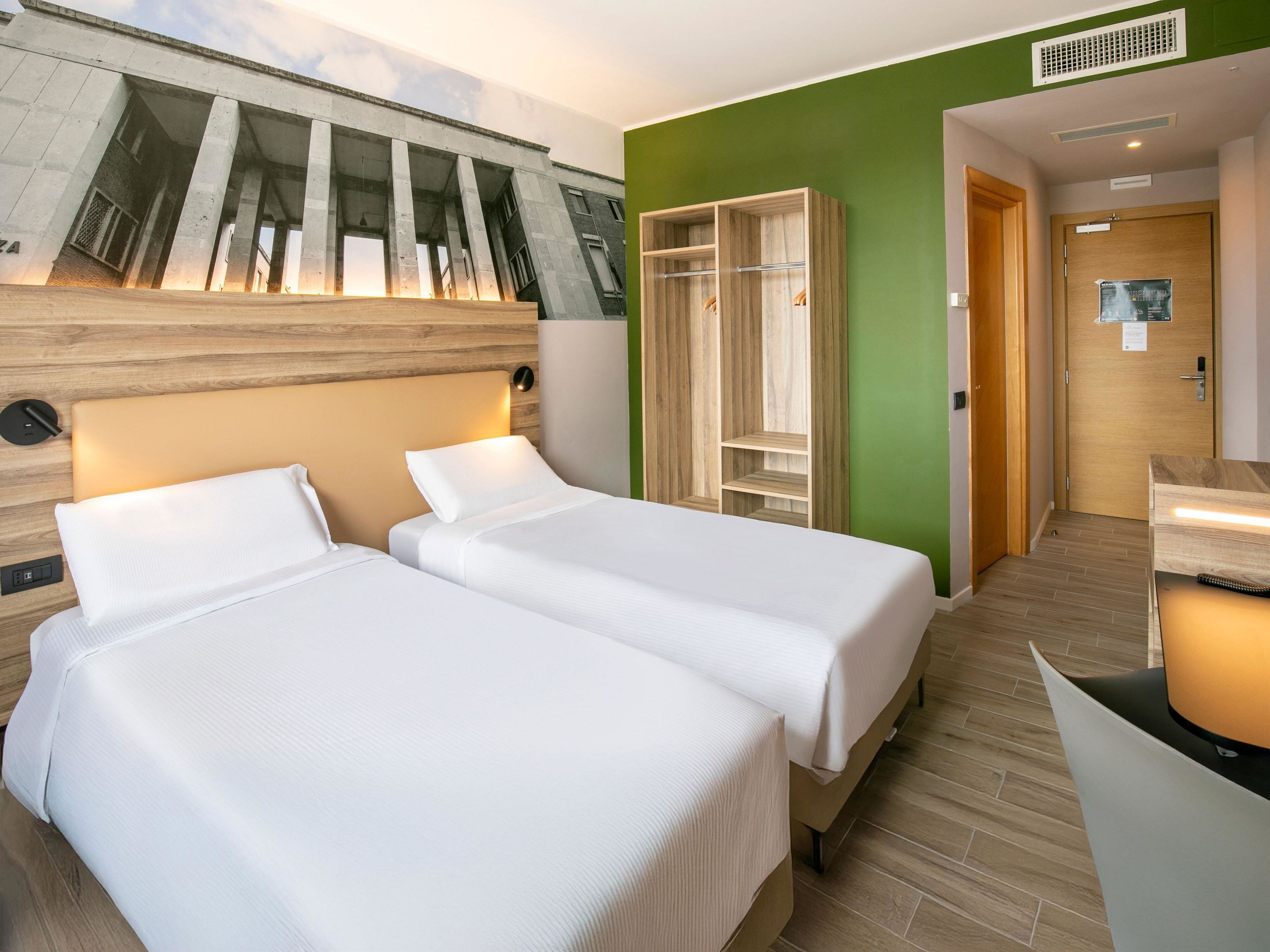 Hotelkamer met twee bedden, een houten hoofdbord, een groene accentmuur en een inbouw kast.