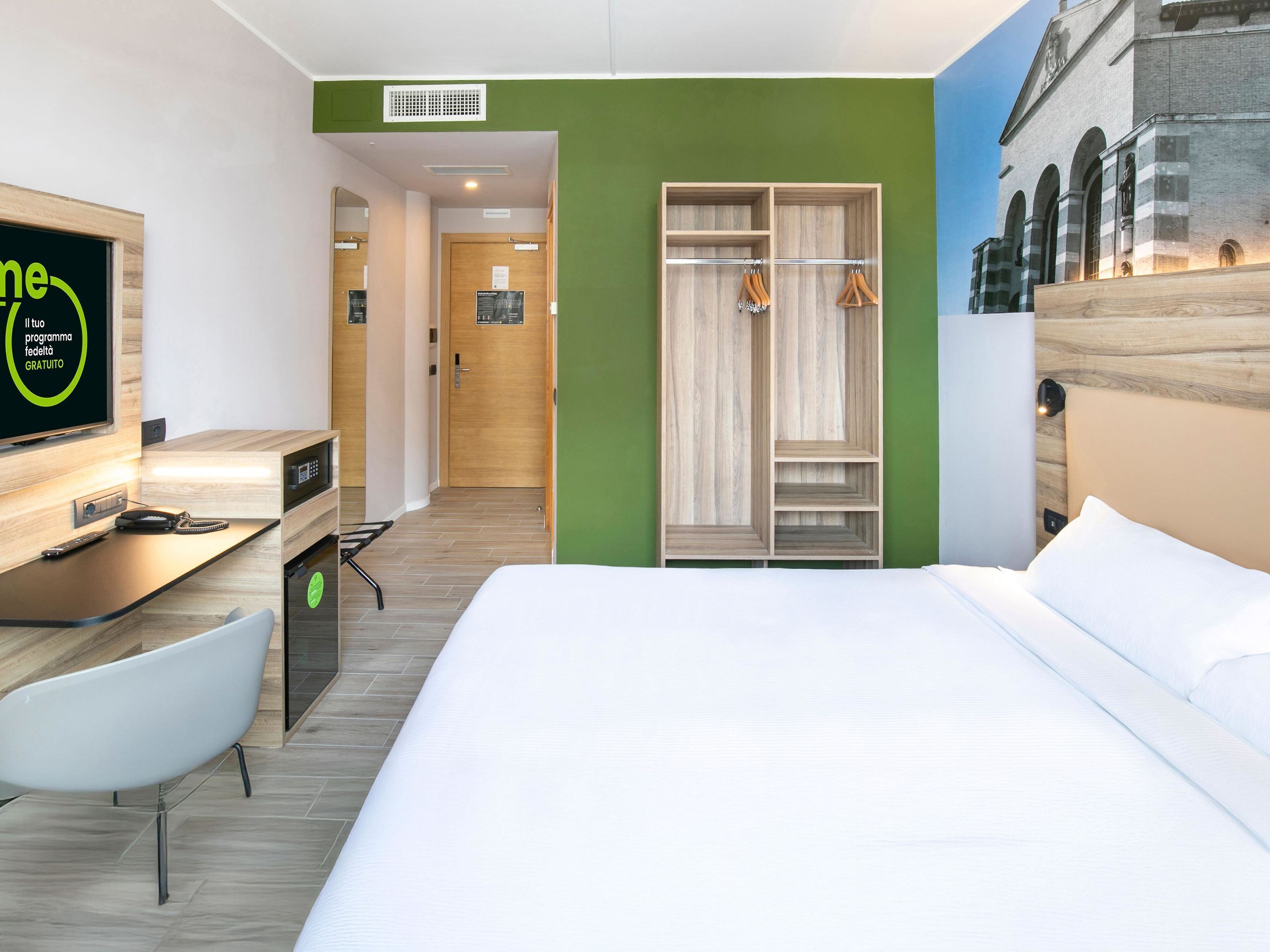 Ein Hotelzimmer mit Bett, Schreibtisch inklusive Computer, Stuhl, Kleiderschrank und Fernseher, der ein Programm zeigt.