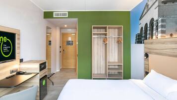 Ein Hotelzimmer mit Bett, Schreibtisch inklusive Computer, Stuhl, Kleiderschrank und Fernseher, der ein Programm zeigt.