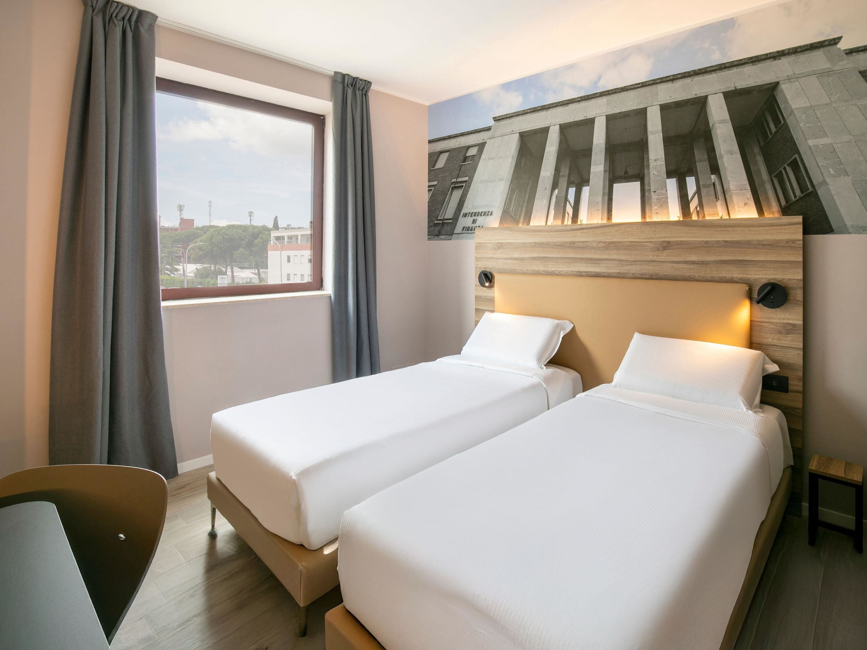 A hotel room with two single beds, a large window, and a view of the cityscape.