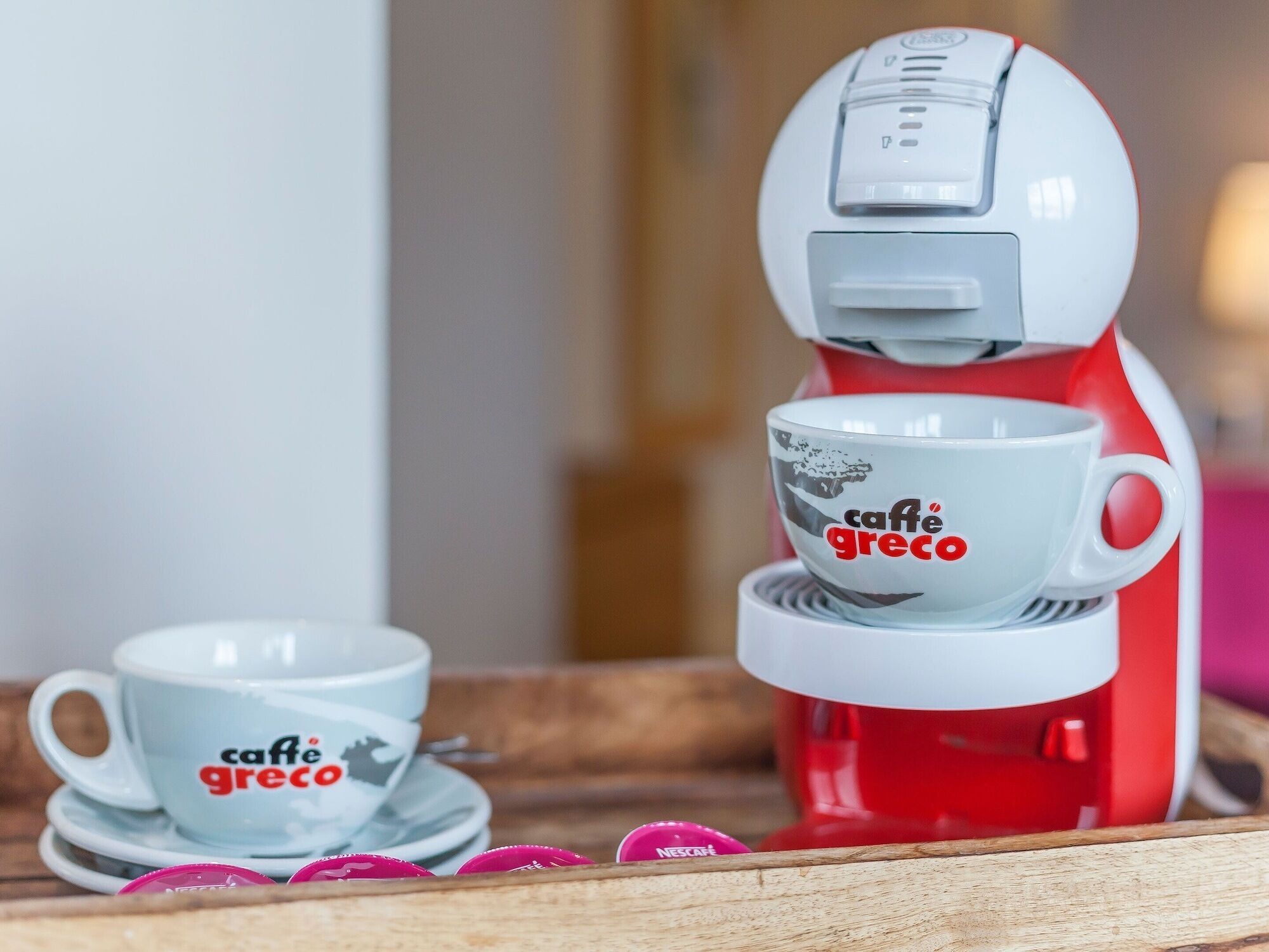 Una cafetera con una taza y platillo etiquetados como 'caffe greco' y una bandeja con cápsulas de café.