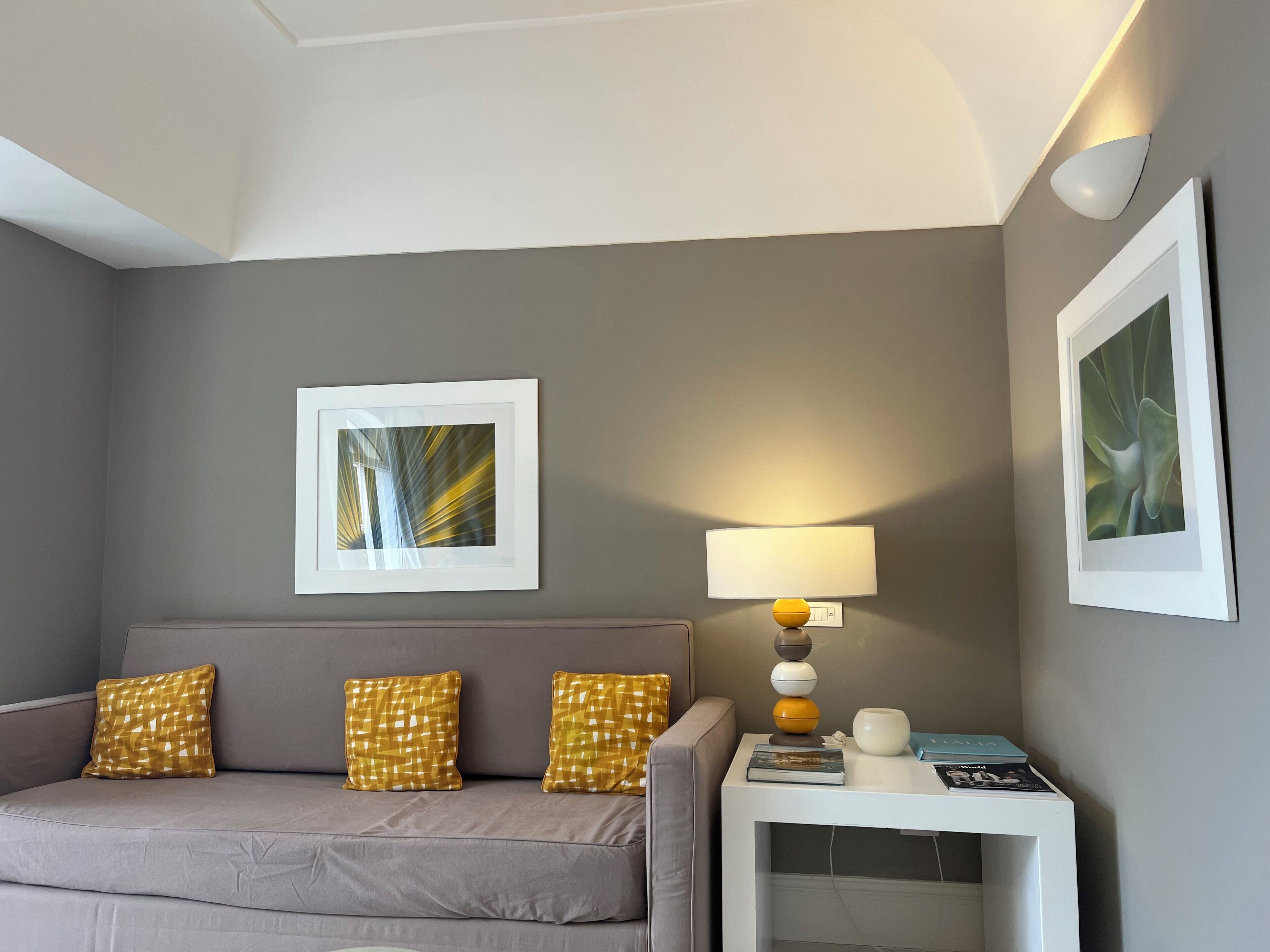 Habitación de hotel moderna con un sofá gris, dos almohadas amarillas estampadas, una mesita de noche blanca con lámpara y cuadros en las paredes.
