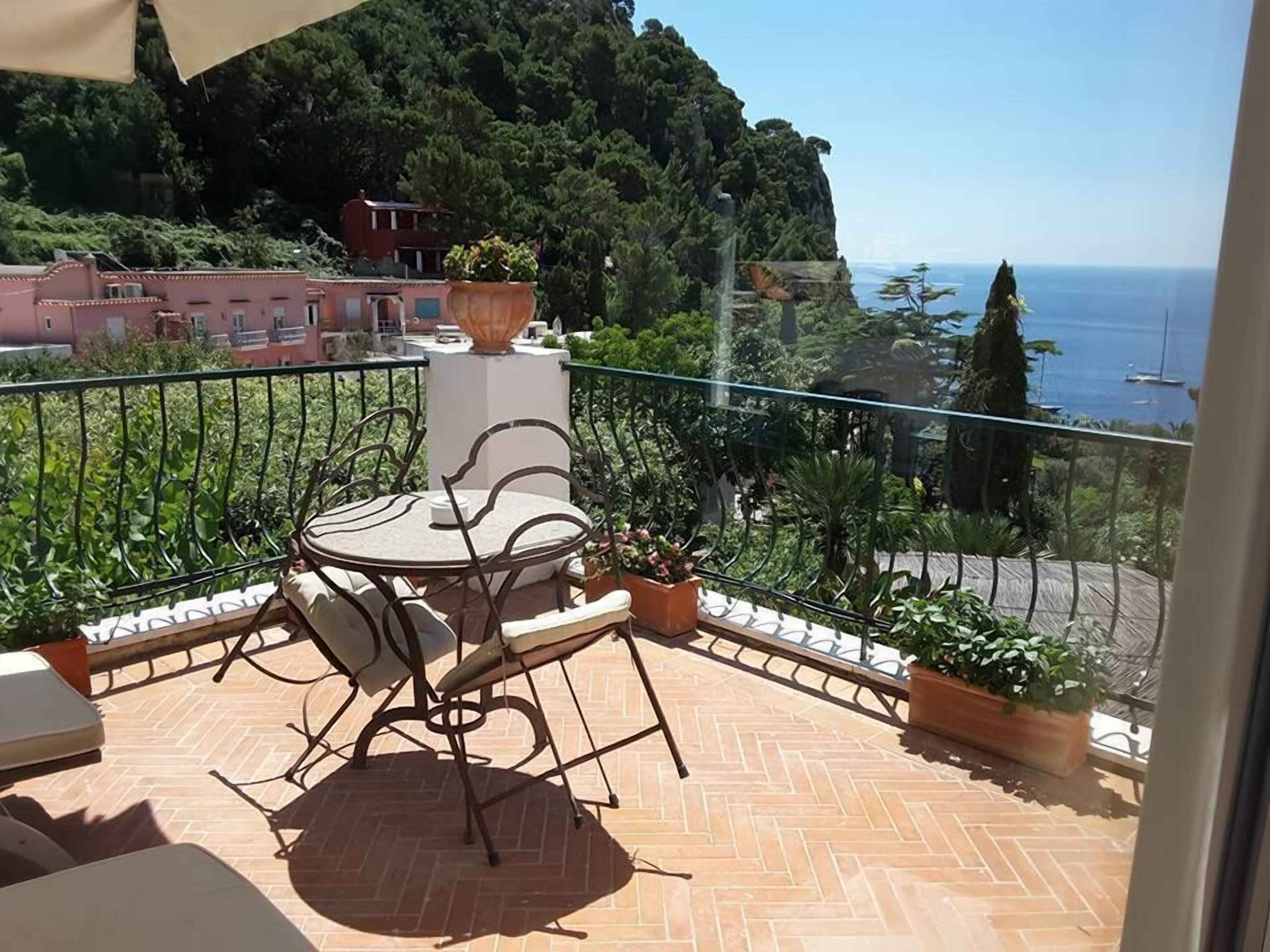 A balcony with a table and chairs, potted plants, and a view of the sea and hills.