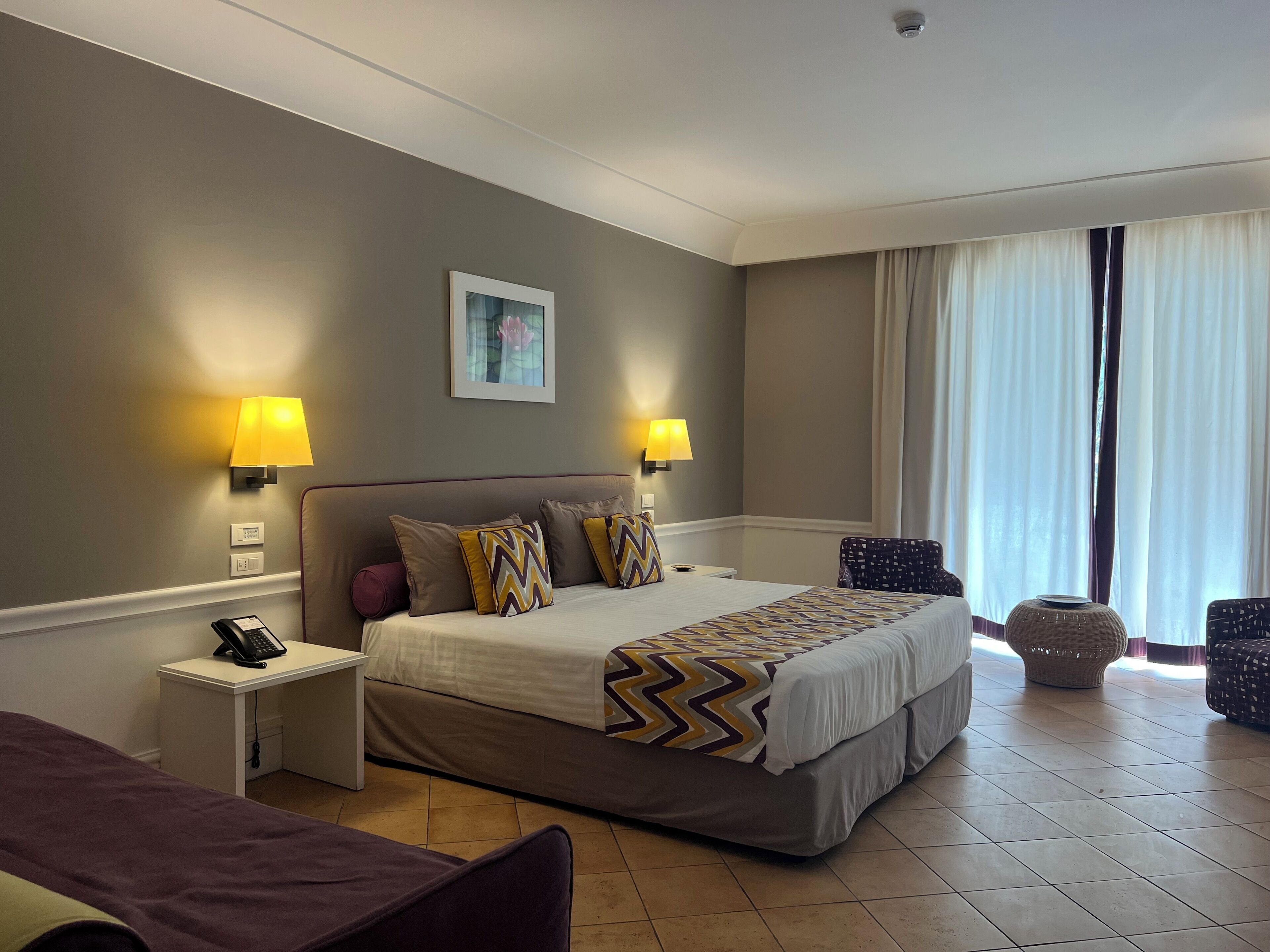 A hotel room with a large bed, two bedside lamps, a small table with a phone, a chair, and a window with curtains.