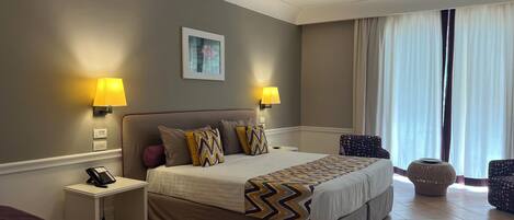 A hotel room with a large bed, two bedside lamps, a small table with a phone, a chair, and a window with curtains.